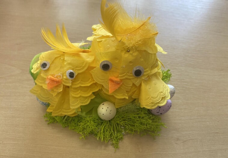Przestrzenna praca plastyczna – dwa kurczaki w kształcie jajka mające piórka z żółtych serwetek, stoją na zielonej trawce wśród pisanek.