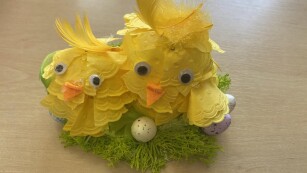 Przestrzenna praca plastyczna – dwa kurczaki w kształcie jajka mające piórka z żółtych serwetek, stoją na zielonej trawce wśród pisanek.