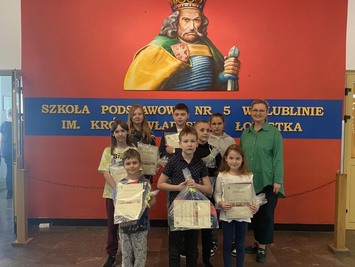Cztery dziewczynki i czterech chłopców wraz z wicedyrektor szkoły stoją z nagrodami na tle muralu przedstawiającego Króla Władysława Łokietka.