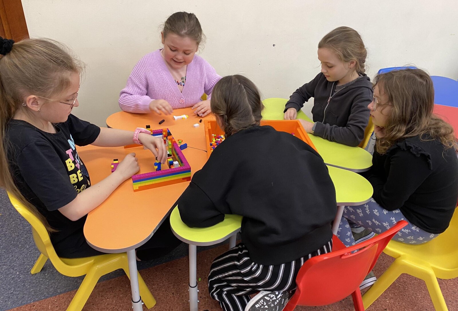 Pięć dziewczynek siedzi na krzesełkach przy stoliku i bawi się klockami typu lego. Konstruuje z nich ludziki oraz dom.