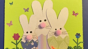 5. Kartka wielkanocna – trzy króliki wycięte z papieru, wyłaniające się z kolorowych skorupek jajek. Obok kwiaty.